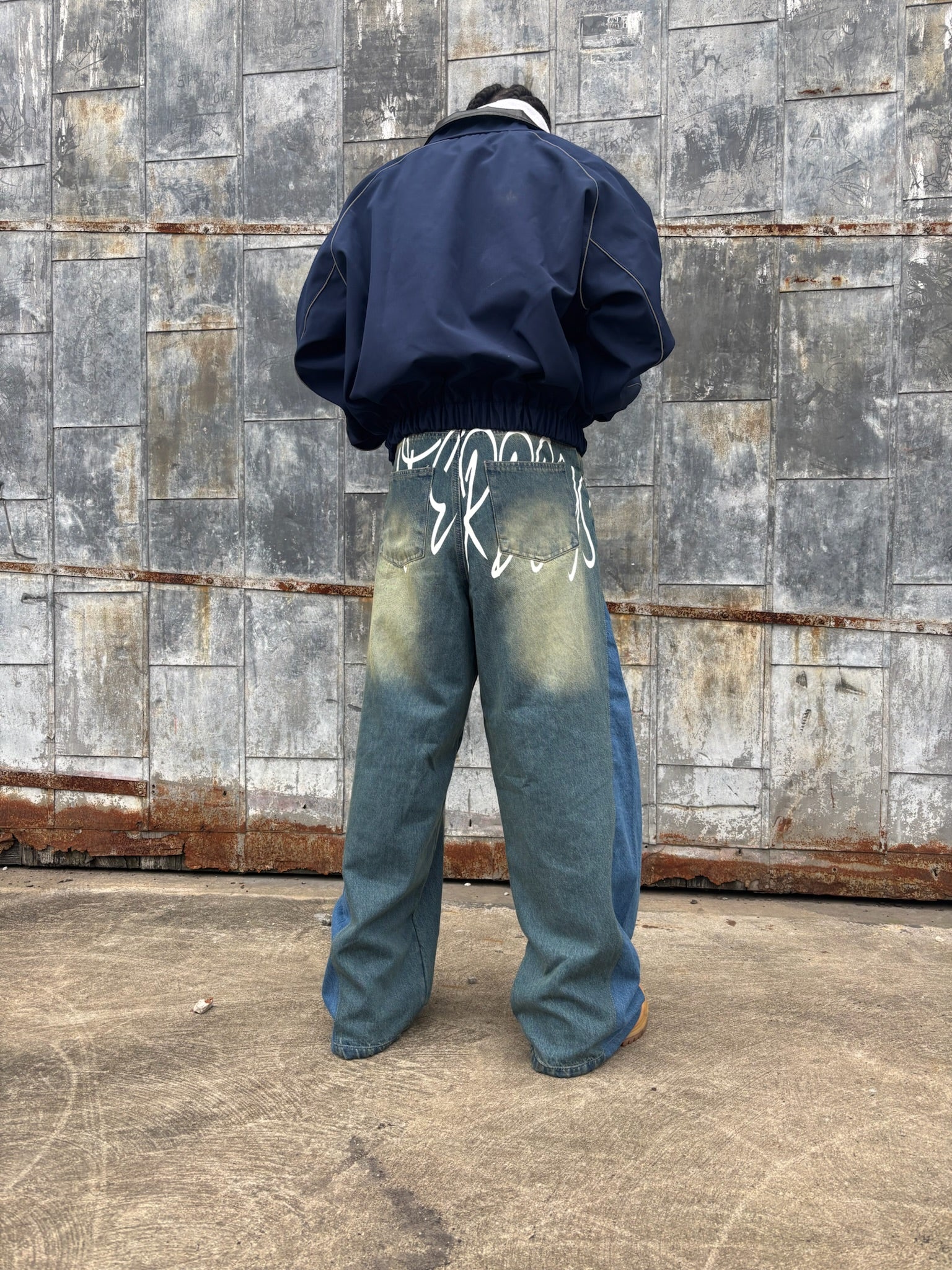 Person wearing a navy jacket and blue jeans with white paint splashes, standing against a textured wall.