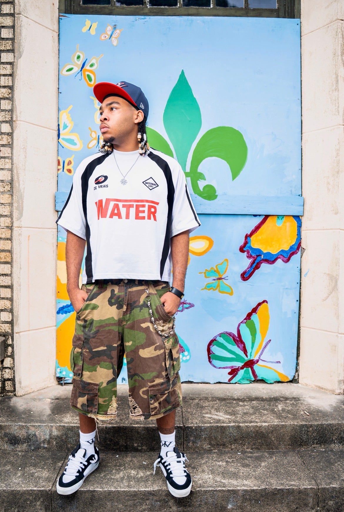 Person wearing a white t-shirt with 'WATER' printed on it, camouflage shorts, and black and white sneakers, standing in front of a colorful mural.