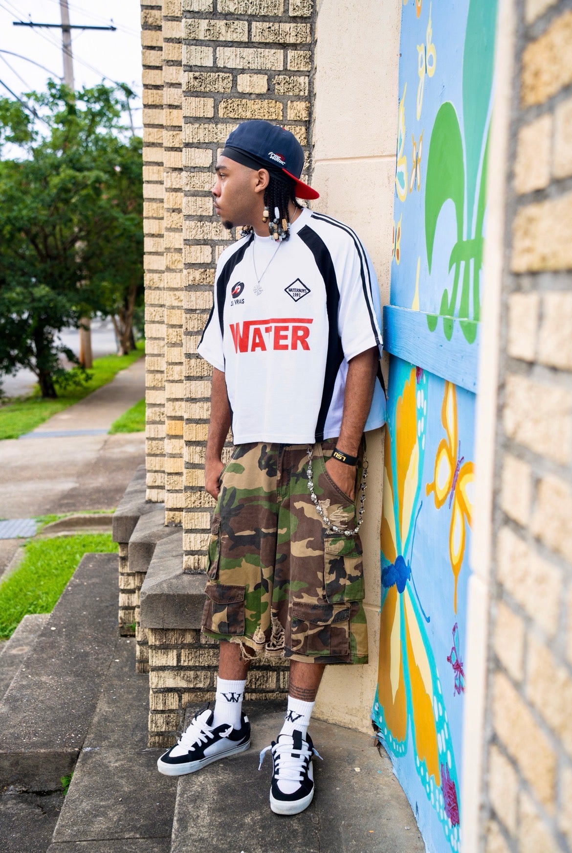 Person wearing a white t-shirt with red text, camouflage shorts, and black and white sneakers standing against a colorful mural.